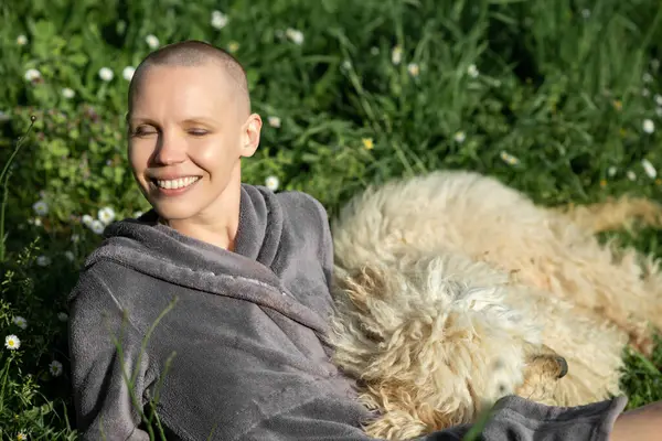 Joyful contented bald woman with a dog is lying on the green grass on a sunny day. Joy of life concept. Home pet.