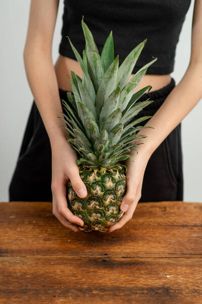 Close-up of unrecognizable person holding a ripe pineapple, set against a neutral background on a rustic wood surface.