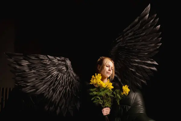 A woman with black angel wings stands in contrast to the dark background, representing the duality of good and evil. The yellow flowers in her hands represent the struggle between good and evil.