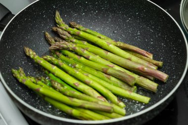 Bir tava kuşkonmaz ocakta pişiyor. Kuşkonmaz yeşildir ve birkaç kahverengi ucu vardır.