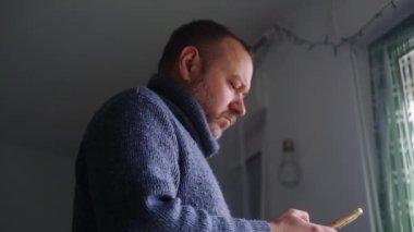 Mavi kazaklı bir adam pencerenin önünde oturuyor. Telefonunda bir şeye bakıyor.