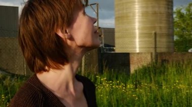 Bir kadın kır çiçeklerinin arasında duruyor, sakin bir an endüstriyel arkaplanda yan yana..