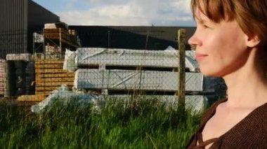 Bir kadın uzun otların arasında huzur içinde duruyor, batan güneş yüzüne sıcak bir parıltı ve arkasında endüstriyel bir manzara bırakıyor..