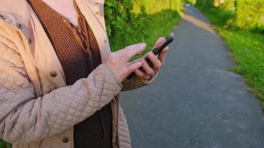 Bir insan telefonunu kontrol ederken parkta bir patika boyunca yürür. Güneş parlıyor ve ağaçlar yeşil.