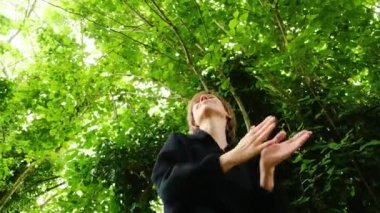 Bir kadın ormanda dikiliyor, gökyüzüne bakıyor. Sahne huzurlu ve sakin, kadın doğayla çevrili ve o anın tadını çıkarıyor.