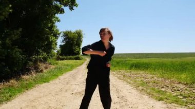 Genç bir adam toprak bir yolda dururken ellerini havaya kaldırarak oynuyor. Sahne huzurlu ve sakin, çocuk dışarıda eğleniyor.