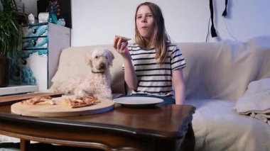 Genç kız, parlak oturma odasındaki rahat koltukta evcil köpekle pizzayı seviyor..