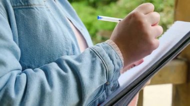 Bir kişi kalemle deftere yazıyor. Defter, üzerinde bir sürü yazı olan bir sayfaya açıktır. Mavi tişörtlü ve defterin üzerinde eli olan kişi.