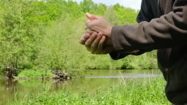 Bir adam çimenli bir arazide bir su kütlesinin yanında duruyor. Kahverengi bir ceket giyiyor ve cebinde bir eli var. Sahne sakin ve sakin, adamın dışarıda eğlendiği bir yer.