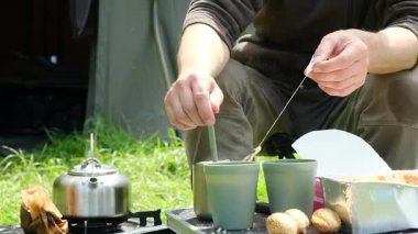 Bir adam elinde beyaz bir nesneyle çimlerin üzerinde oturuyor. Kahverengi bir ceket giyiyor ve siyah bir kapüşonlu giyiyor.