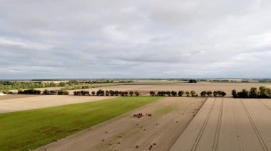 Bulutlu gökyüzünün altında geniş kırsal alanlarda ilerleyen hasat makinelerinin havadan görünüşü.