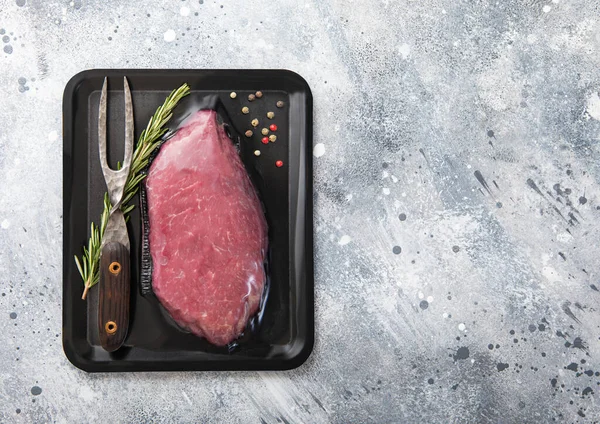 Raw beef steak sirloin fillet sealed in vacuum tray with pepper,rosemary and barbeque fork on light kitchen table background.Space for text