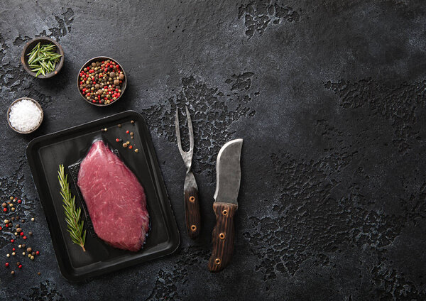 Beef raw fillet steak in vacuum tray with pepper,salt and rosemary on black background with barbeque fork and knife.Top view.