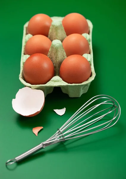 Raw eggs in paper tray with whisk,yolk and shell on green.