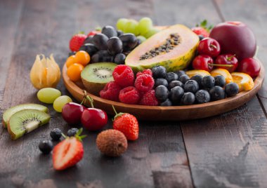 Fresh raw organic summer berries and exotic fruits in round wooden plate on dark wooden kitchen background. Papaya, grapes, nectarine, orange, raspberry, kiwi, strawberry, lychees, cherry. Top view clipart