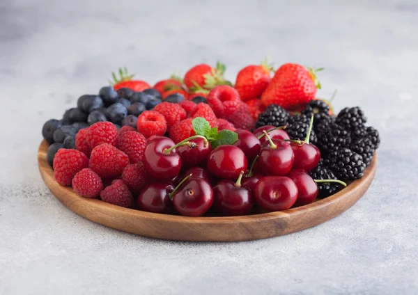 Fresh organic summer berries mix in round wooden tray on light kitchen table background. Raspberries, strawberries, blueberries, blackberries and cherries. Top view