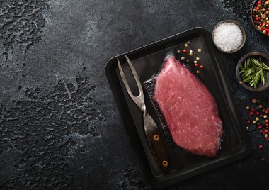 Vacuum tray with fresh raw beef steak sirloin fillet with pepper,rosemary and barbeque fork on black background.Top view.