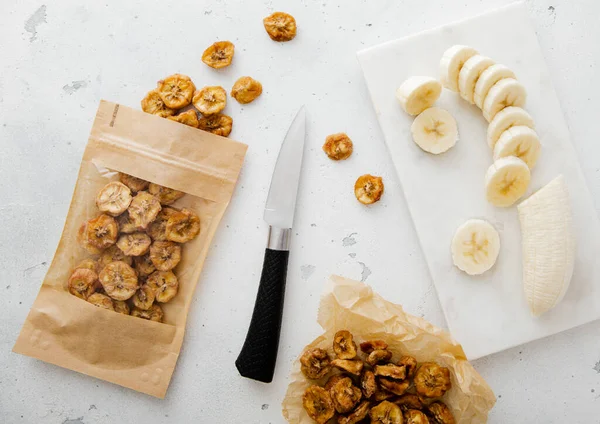 Pack of dried bio banana chip slices with knife and raw banana.