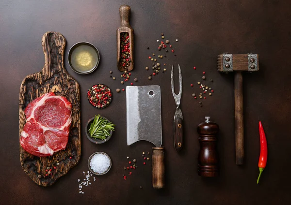 Meat cleavers,tenderizer,fork,knife and other kitchen meat utensils with oil,herbs,salt and pepper on dark background with raw rib eye steak fillet.Top view.
