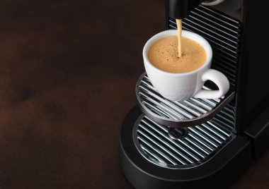 Kahverengi mutfak arka planındaki beyaz fincana ev yapımı espresso makinesinden taze sabah kahvesi dökülüyor..