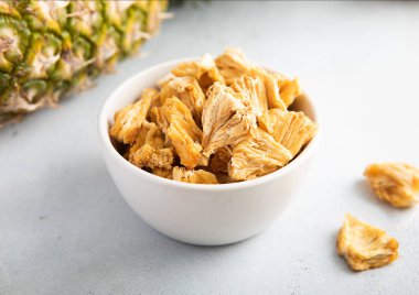 Kurutulmuş ananas parçaları cam kasede taze ananas meyveleriyle birlikte hafif arka planda..