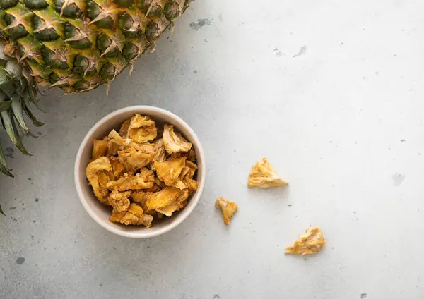Dried pineapple slices chips in glass bowl on light background with ripe pineapple fruit.Top view.