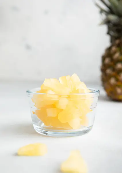 Dried soft sweet pineapple slices in glass bowl with raw pineapple on light table.