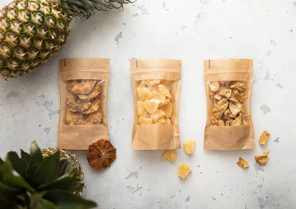 Packages with dried sweet pineapple slices and circles on light background with raw pineapple fruit.