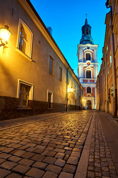 Cobblestone street with baroque bell tower of a historic monastery at night in Poznan, Poland