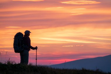 Hiker woman with backpack on the mountain top looking at the scenic sunset sky. Exploring, achievement or active lifestyle concept.