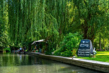 The image depicts a tranquil river landscape, with boats, wooden bridges, and single-family homes along the water. Surrounded by nature and landscape, the scene has a natural and peaceful atmosphere. Lbbenau, Brandenburg, Germany