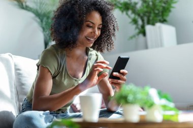 Evdeki kanepede oturup kahve içerken kendine güvenen bir kadının cep telefonunu kullanması..