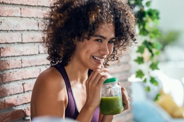Evdeki oturma odasında duran sağlıklı yeşil smoothie içen mutlu bir kadın..