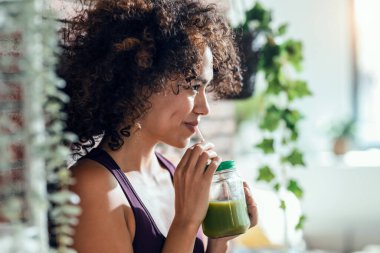 Evdeki oturma odasında duran sağlıklı yeşil smoothie içen mutlu bir kadın..