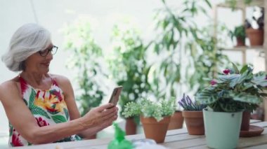 Evde akıllı bir telefonla yeşil bitkilerin ve çiçeklerin fotoğrafını çeken mutlu yaşlı bir kadının videosu..