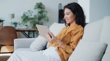 Evdeki kanepede otururken dijital tabletini okuyan güzel bir kadının videosu..