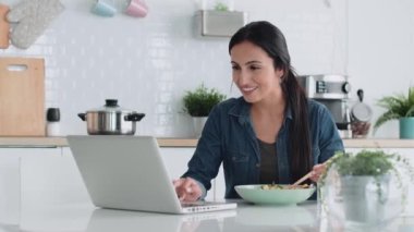 Evdeki mutfakta yemek yerken bilgisayarla çalışan neşeli bir kadının videosu..