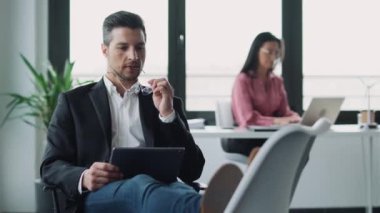 Ofisteki bir sandalyede rahatça otururken dijital tabletle çalışan zarif iş adamının videosu..