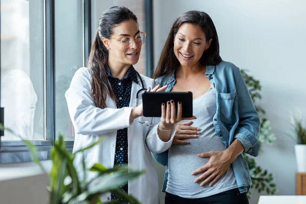 Shot of female gynecologist doctor showing to pregnant woman ultrasound scan baby with digital tablet in medical consultation.