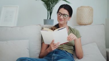 Evde kanepede oturan kendine güvenen bir kadının kitap okuduğu video..