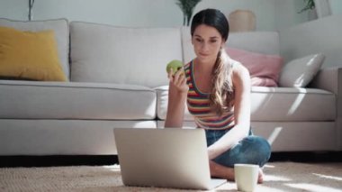 Kendine güvenen bir kadının evinde yerde oturmuş elma yerken bilgisayarını kullanmasının videosu..