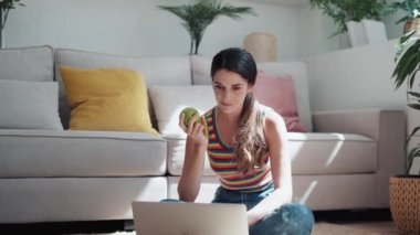 Kendine güvenen bir kadının evinde yerde oturmuş elma yerken bilgisayarını kullanmasının videosu..