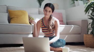 Kendine güvenen bir kadının evinde yerde oturmuş elma yerken bilgisayarını kullanmasının videosu..