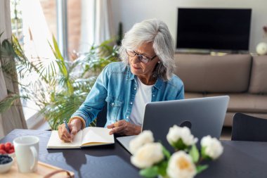 Shot of beautiful senior woman working with laptop while checking some documents in the kitchen at home. 