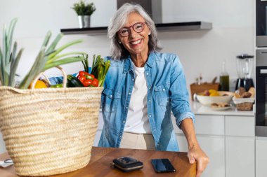 Shot of happy mature woman holding a paper bag full of groceries from the supermarket in the kitchen at home.