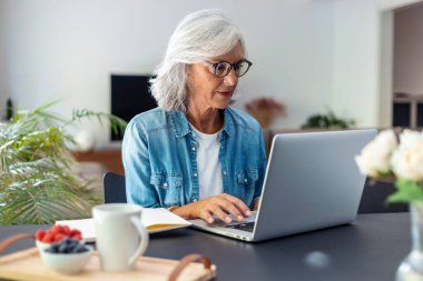 Shot of beautiful senior woman working with laptop while checking some documents in the kitchen at home. 