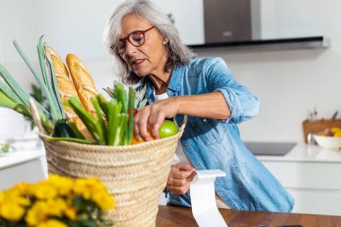 Portrait of lovely mature woman standing at home table holding grocery receipt discussing for rising prices. 