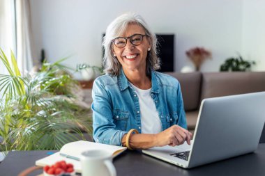 Shot of beautiful senior woman working with laptop while checking some documents in the kitchen at home. 