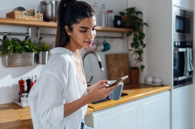 Shot of beautiful woman using her mobile phone while drinking a cup of coffee in the kitchen at home.