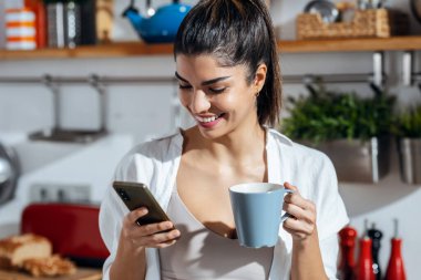 Shot of beautiful woman using her mobile phone while drinking a cup of coffee in the kitchen at home.